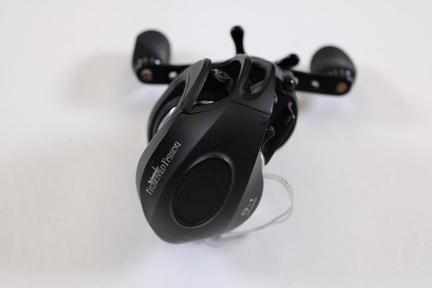 A used Fitzgerald Stunner 6.3:1 LH casting reel in excellent condition, with “PCASTING FISHING” on its side, sits on a white surface. This compact black reel with dual handles is perfect for rod and reel trade-in upgrades.