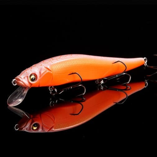 A vibrant orange Megabass ITO Vision 110 (SP-C) Secret V-Ore jerkbait with a textured body, three treble hooks, and a clear lip is displayed on a glossy black background with its reflection below.