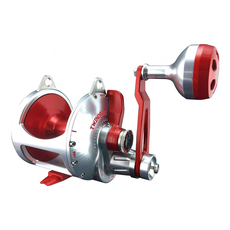 A silver and red fishing reel with a large, round handle. The reel has a shiny finish, multiple mounting points, and intricate detailing, showcasing a robust design for deep-sea or heavy-duty fishing.