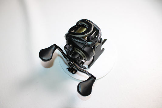 A Lew's Laser Pro LP1SHB used casting reel in fair condition, featuring dual handles, is shown from above on a white circular surface against a plain white background.