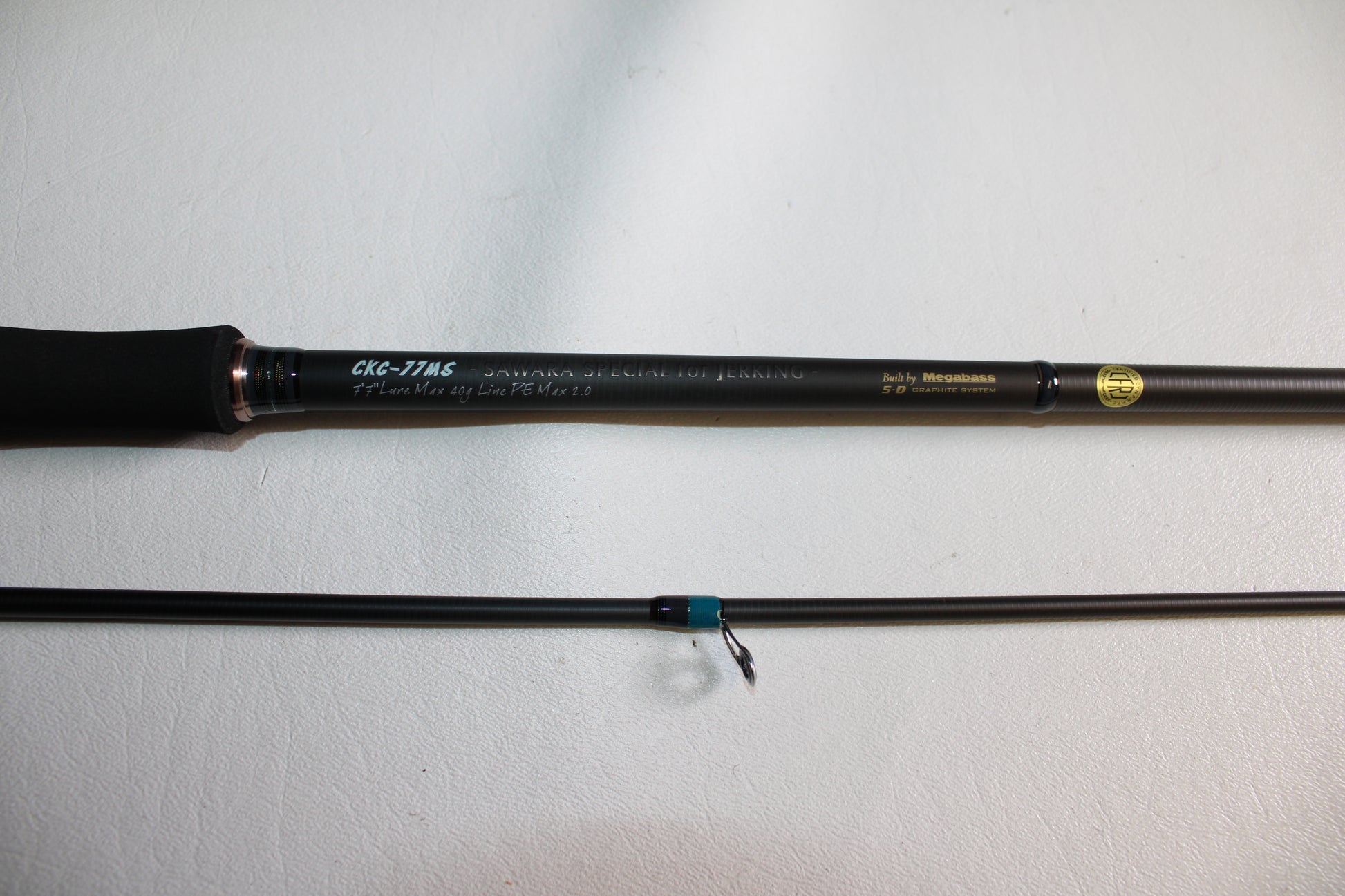 A close-up of a Megabass Cookai Gulf CKG77MS 2 Piece Used Spinning Rod in excellent condition, showing its handle and specifications on a white surface, with another rod section parallel below.