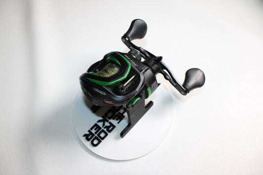 A Lew's Mach Speed Spool MS1SHL casting reel in black and green is shown on a white circular stand with black text. The used reel features dual handles, a green fishing line spool, and a plain white background.