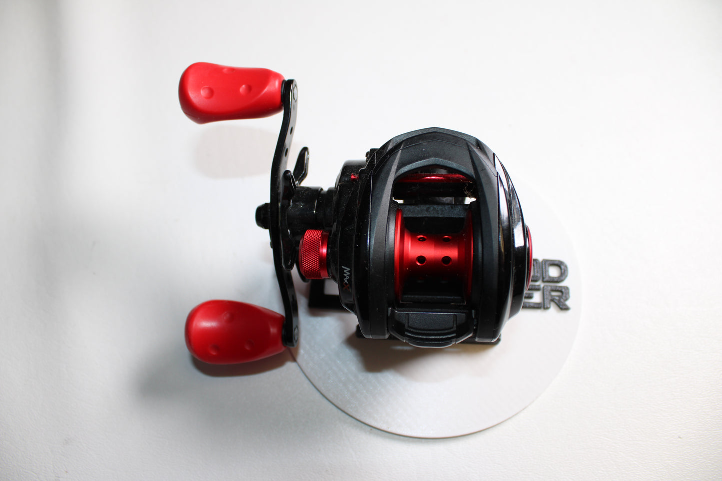 An Abu Garcia MAX4X-L 6.4:1 LH baitcasting reel in black and red with two red handles, shown on a white surface with a partially visible round white sticker; used, in good condition.