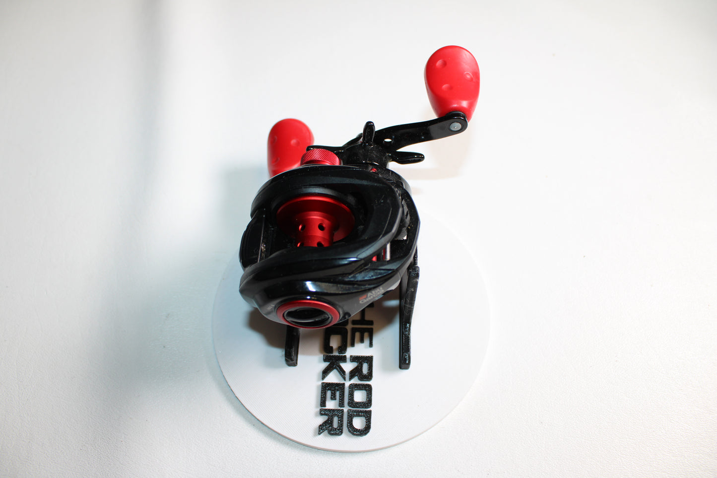 An Abu Garcia MAX4X-L 6.4:1 gear ratio left-hand used casting reel in black and red, with two handles, shown on a white circular base with partially visible "THE ROD LOCKER" text. Good condition.