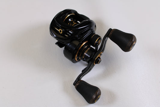 A Lew's Lite MgF LFS 7.5:1 LH used casting reel in good condition, featuring dual handles and a black-and-gold design, rests on a white surface. Trade it in for store credit with our Rod and Reel Trade-In Program.
