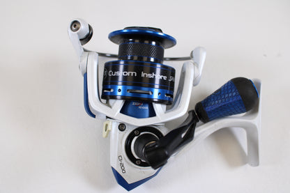 A close-up of a Lew's Custom Inshore CI-200 6.2:1 used spinning reel in good condition, featuring a blue and silver body with a black and blue handle, set against a plain white background.