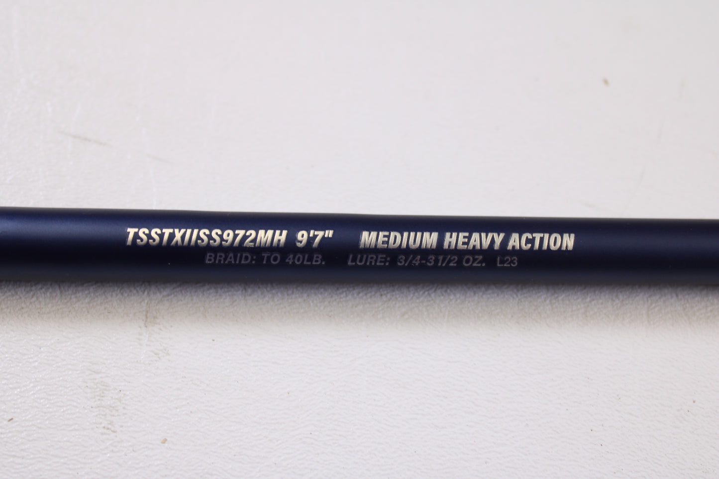 A close-up of a Tsunami SaltX Surf TSSTXIISS771M 7'7" Medium Used Spinning Rod—part of our Rod and Reel Trade-In Program—shows identifying text on the dark rod against a white background. Brand: Tsunami. Condition: Excellent.