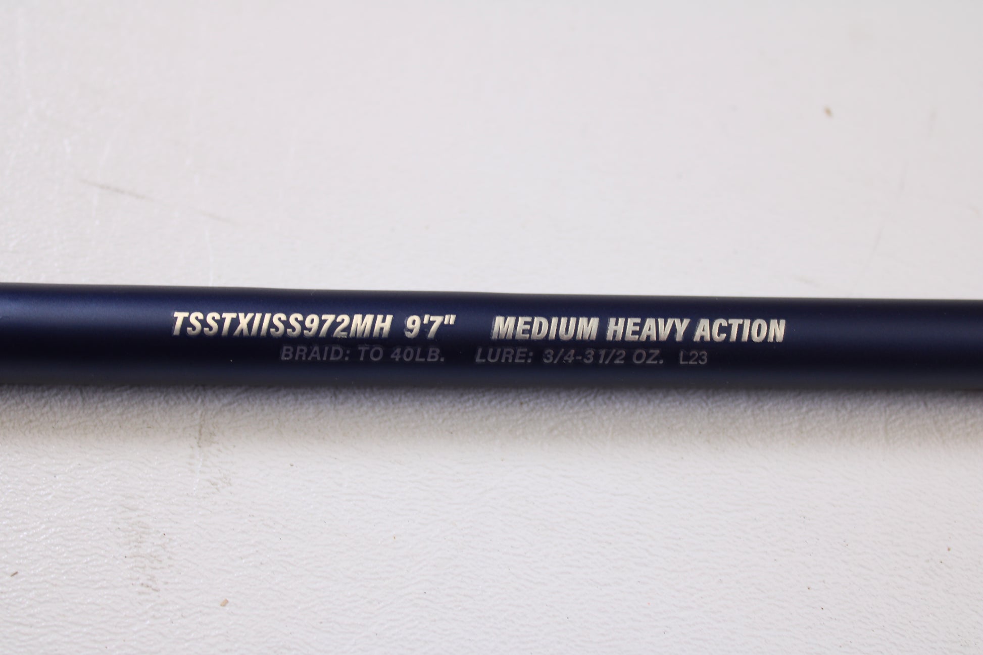 A close-up of a Tsunami SaltX Surf TSSTXIISS771M 7'7" Medium Used Spinning Rod—part of our Rod and Reel Trade-In Program—shows identifying text on the dark rod against a white background. Brand: Tsunami. Condition: Excellent.