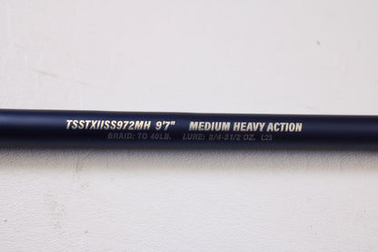 A close-up of a Tsunami SaltX Surf TSSTXIISS771M 7'7" Medium Used Spinning Rod—part of our Rod and Reel Trade-In Program—shows identifying text on the dark rod against a white background. Brand: Tsunami. Condition: Excellent.