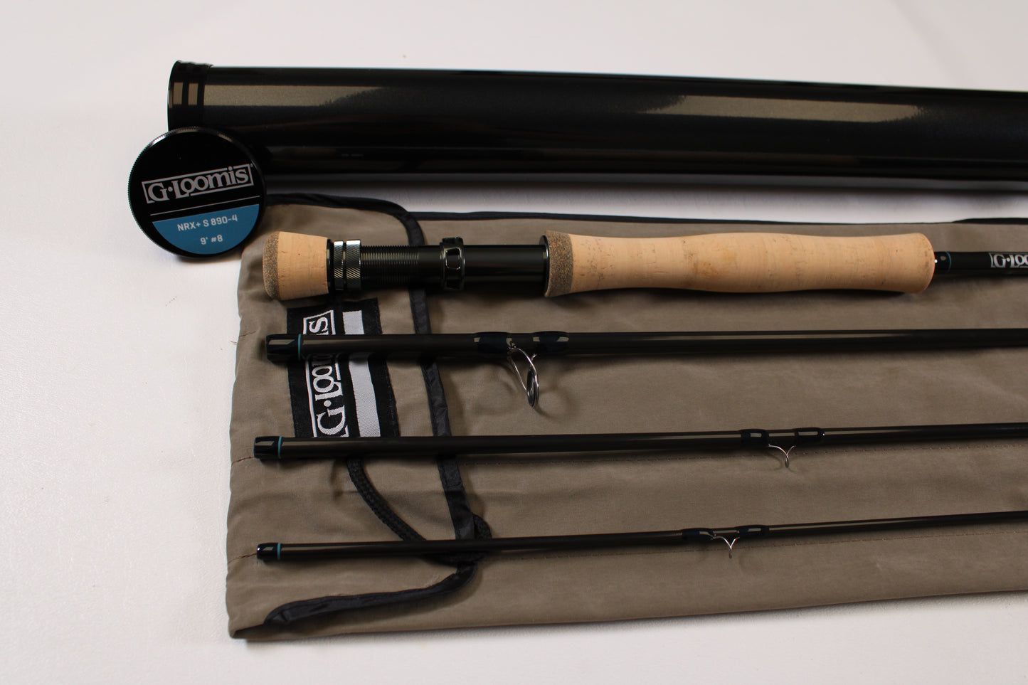 A G. Loomis NRX+ S 890-4 used fly rod in mint condition, black with a cork handle, is disassembled into four pieces on a gray cloth case beside a black rod tube and a round "G. Loomis" container.