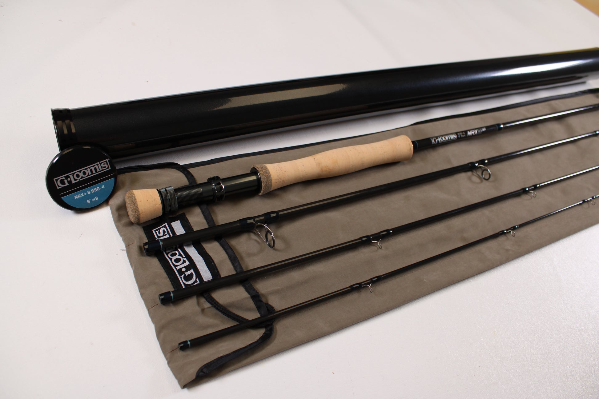 A G. Loomis NRX+ S 890-4 used fly rod in mint condition with a cork handle, shown disassembled on a cloth case beside a black tube and round "G. Loomis" container.