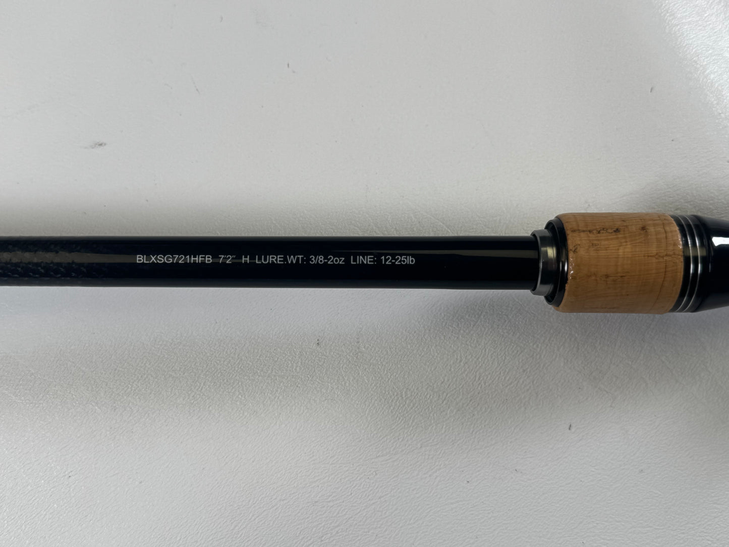 Close-up of a Daiwa Daiw BLX BLXSG721HFB 7'2" Heavy used casting rod handle with "BLKSG721HFB-7'2"-H LURE. WT: 3/8-2oz LINE: 12-25lb" text. The thick cork grip has a glossy black finish. Trade in used gear for store credit through our Rod and Reel Trade-In Program!.