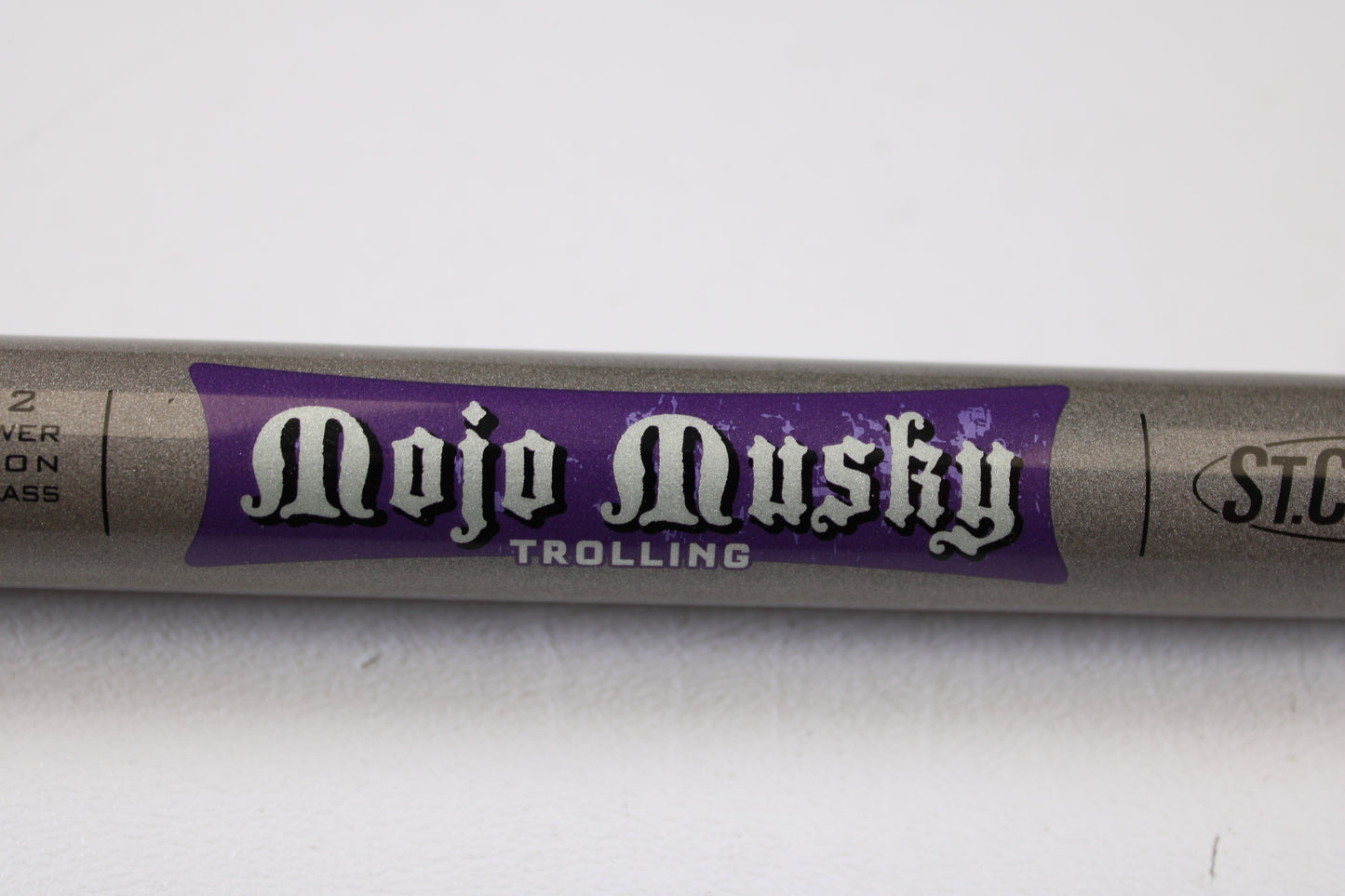 A close-up of a St. Croix Mojo Musky Trolling MMT90XHM2 9'0" XH used casting rod in mint condition, featuring stylized white text on purple, promotes our Rod and Reel Trade-In Program for store credit. The silver-gray rod is shown on a white surface.