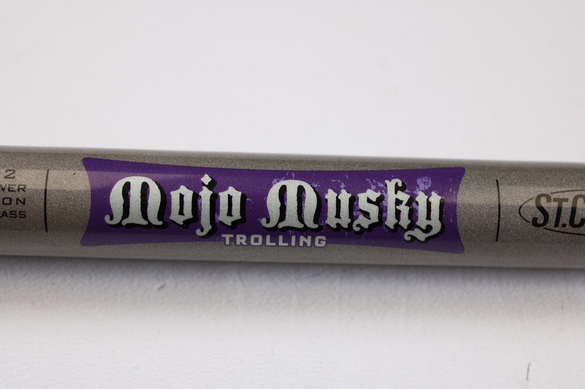 A close-up of a St. Croix Mojo Musky Trolling MMT90XHM2 9'0" XH used casting rod in mint condition, featuring stylized white text on purple, promotes our Rod and Reel Trade-In Program for store credit. The silver-gray rod is shown on a white surface.