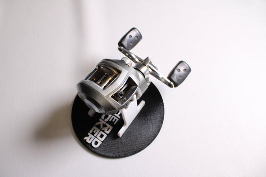 A close-up of an Abu Garcia Revo S-L Left Hand used casting reel in fair condition, displayed on a white surface and mounted on a black circular stand with white lettering.