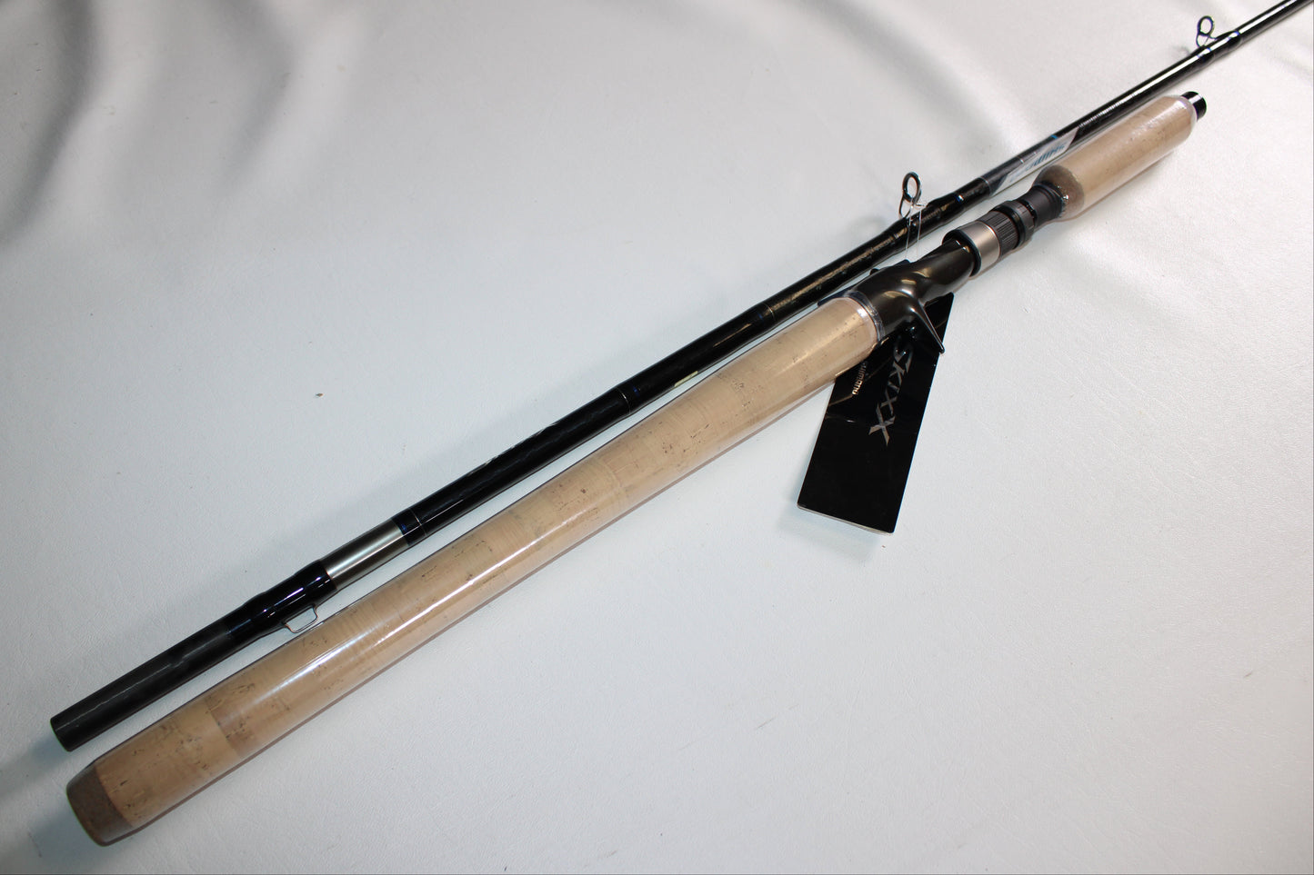 The Shimano SKIXX SKC86XH Used Casting Rod, in mint condition, features a cork handle and black shaft. Shown diagonally on a white surface, it has a black tag attached near the reel seat.