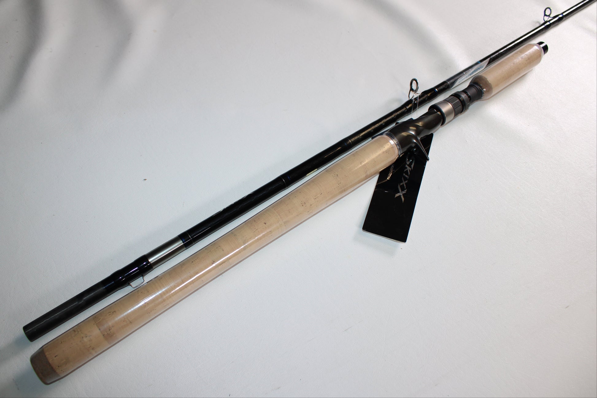 The Shimano SKIXX SKC86XH Used Casting Rod, in mint condition, features a cork handle and black shaft. Shown diagonally on a white surface, it has a black tag attached near the reel seat.