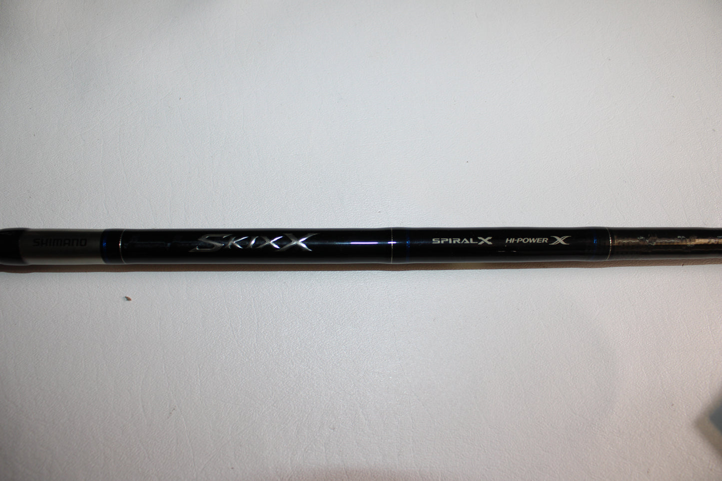 A close-up of the Shimano SKIXX SKC86XH Used Casting Rod in mint condition, showing "Shimano," "SKIXX," "Spiral X," and "Hi-Power X" on the black rod, placed on a white surface.