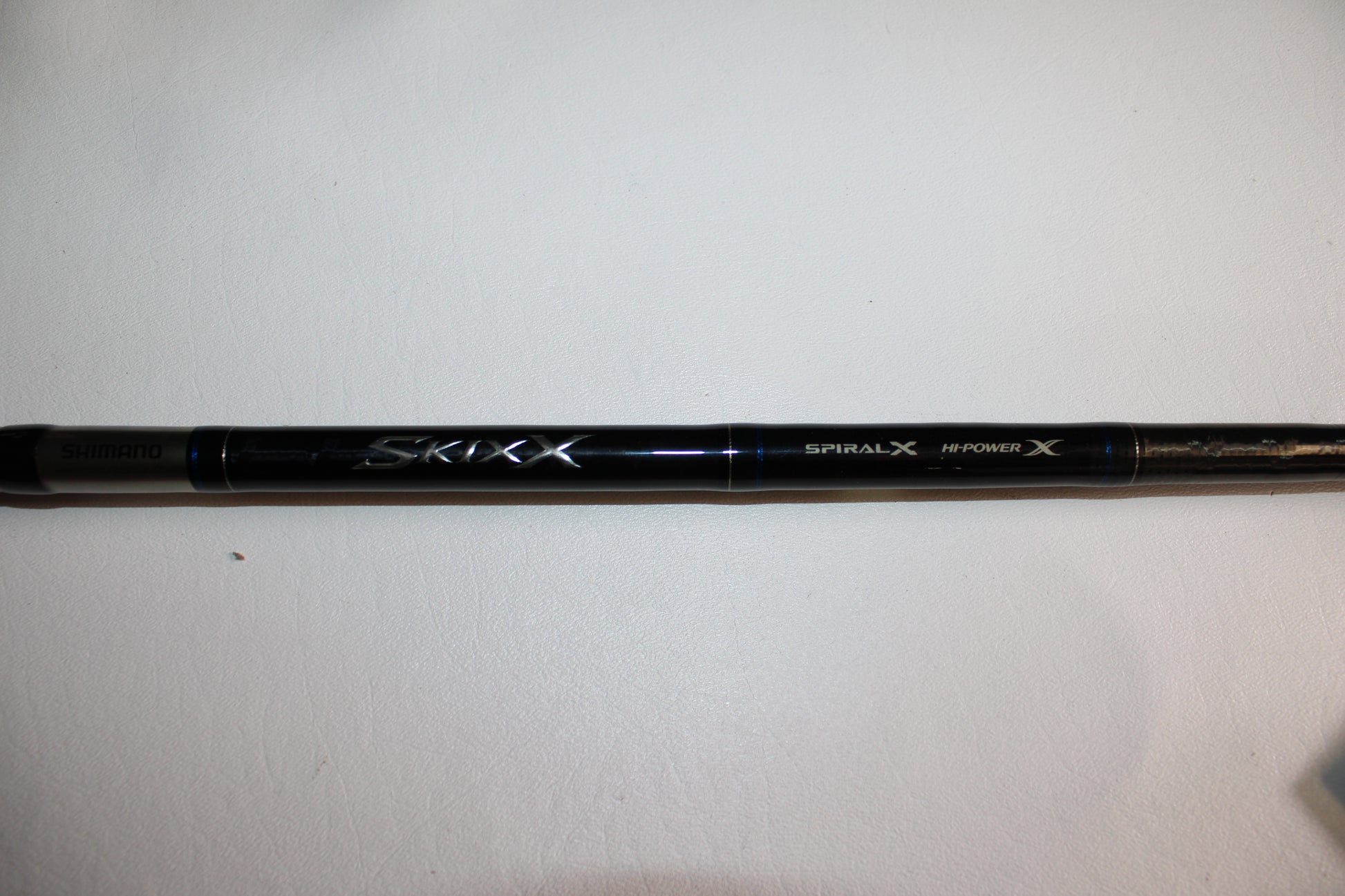 A close-up of the Shimano SKIXX SKC86XH Used Casting Rod in mint condition, showing "Shimano," "SKIXX," "Spiral X," and "Hi-Power X" on the black rod, placed on a white surface.