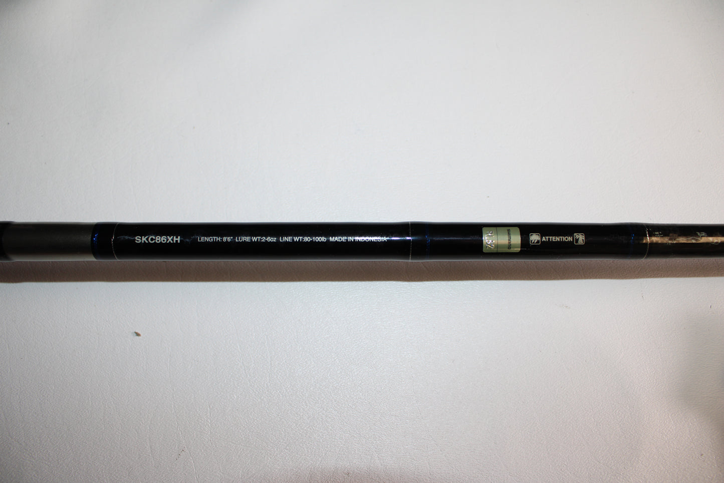 A close-up of the Shimano SKIXX SKC86XH Used Casting Rod in mint condition, with the model number and specs printed in white, plus two small green and gold labels, lying on a white surface.