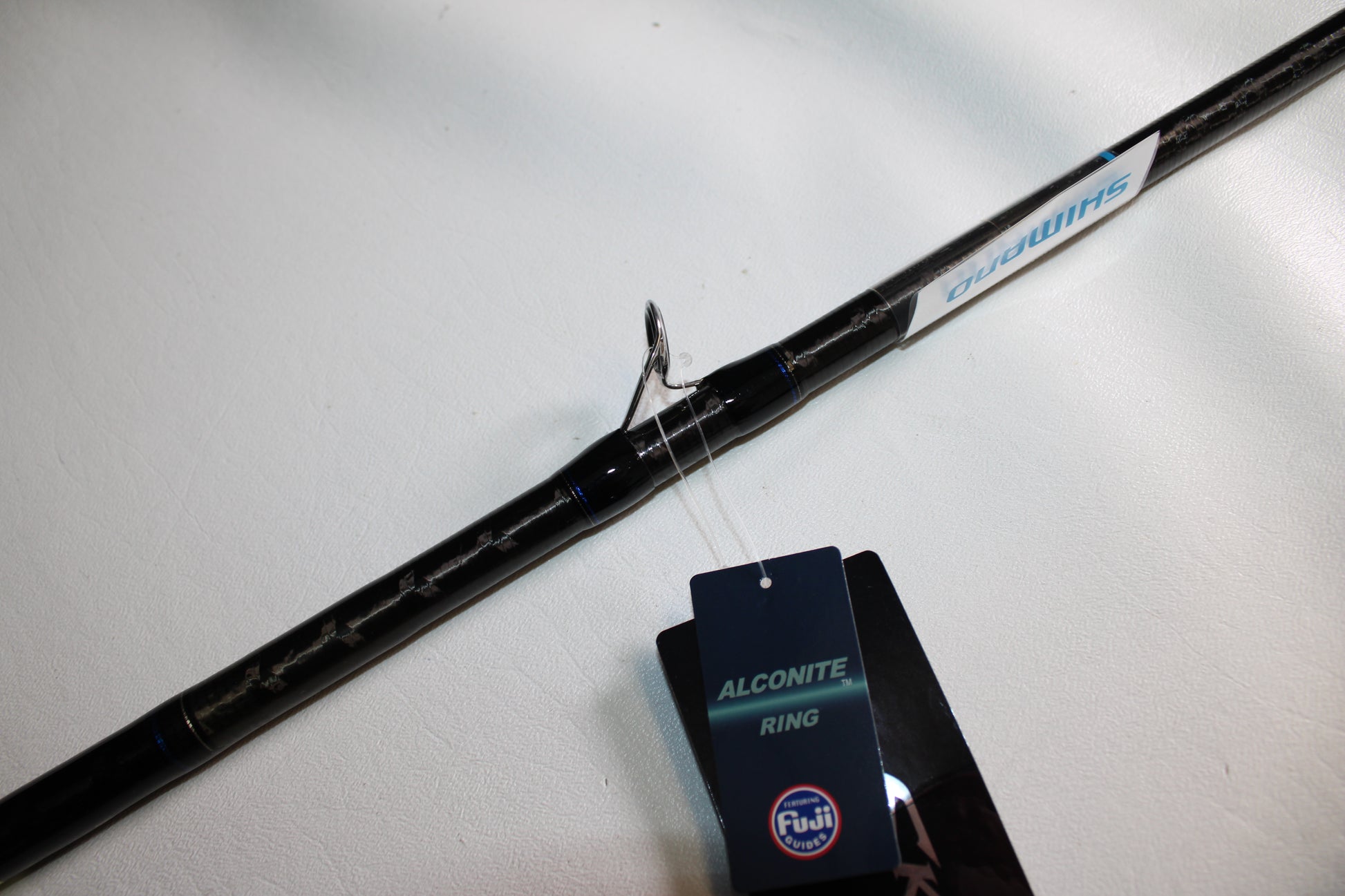 A close-up of the Shimano SKIXX SKC86XH Used Casting Rod in mint condition, featuring the Shimano brand and an "Alconite Ring" tag by Fuji, displayed on a white surface.