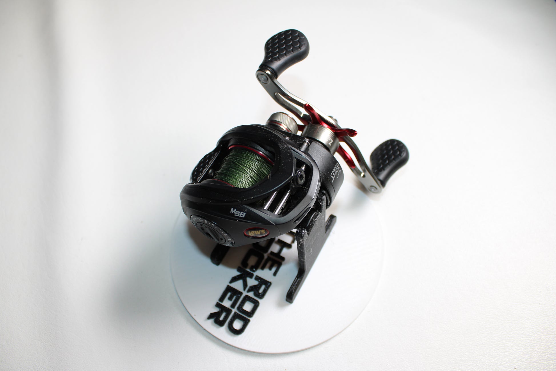 A Lew's Tournament MP TS1SHMPL 7.5:1 used casting reel in excellent condition, shown with green line on a white "THE ROD ROCKER" plate. Features dual textured handles. Upgrade with our Rod and Reel Trade-In Program for store credit!.