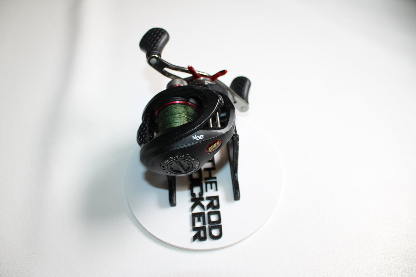A Lew's Tournament MP TS1SHMPL 7.5:1 casting reel in excellent condition is shown on a white stand labeled "THE ROD RACKER." Its handles and dial stand out, making it ideal for our Rod and Reel Trade-In Program.