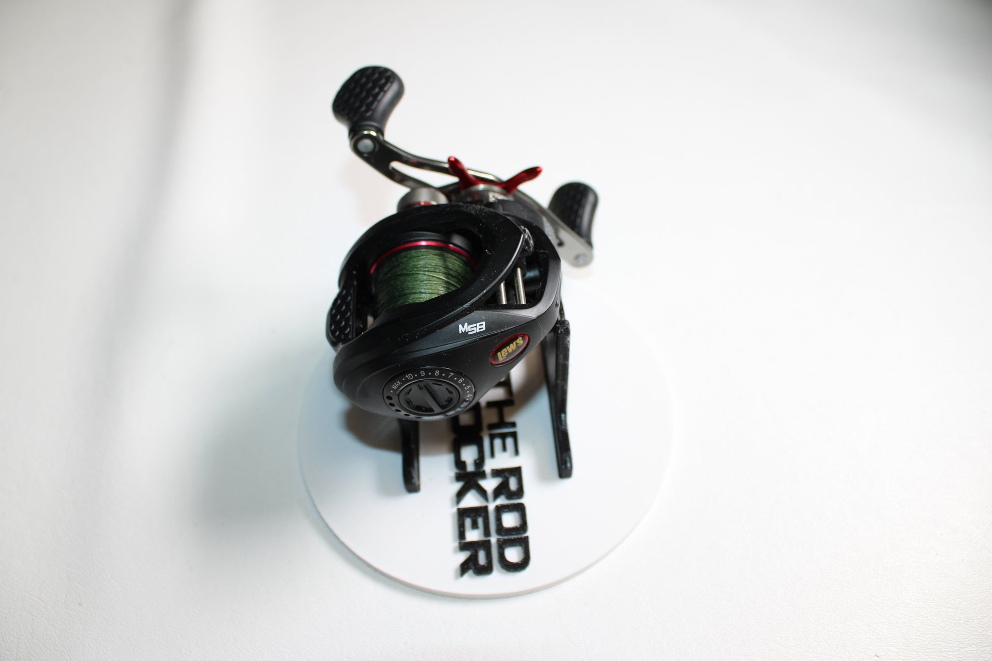 A Lew's Tournament MP TS1SHMPL 7.5:1 casting reel in excellent condition is shown on a white stand labeled "THE ROD RACKER." Its handles and dial stand out, making it ideal for our Rod and Reel Trade-In Program.