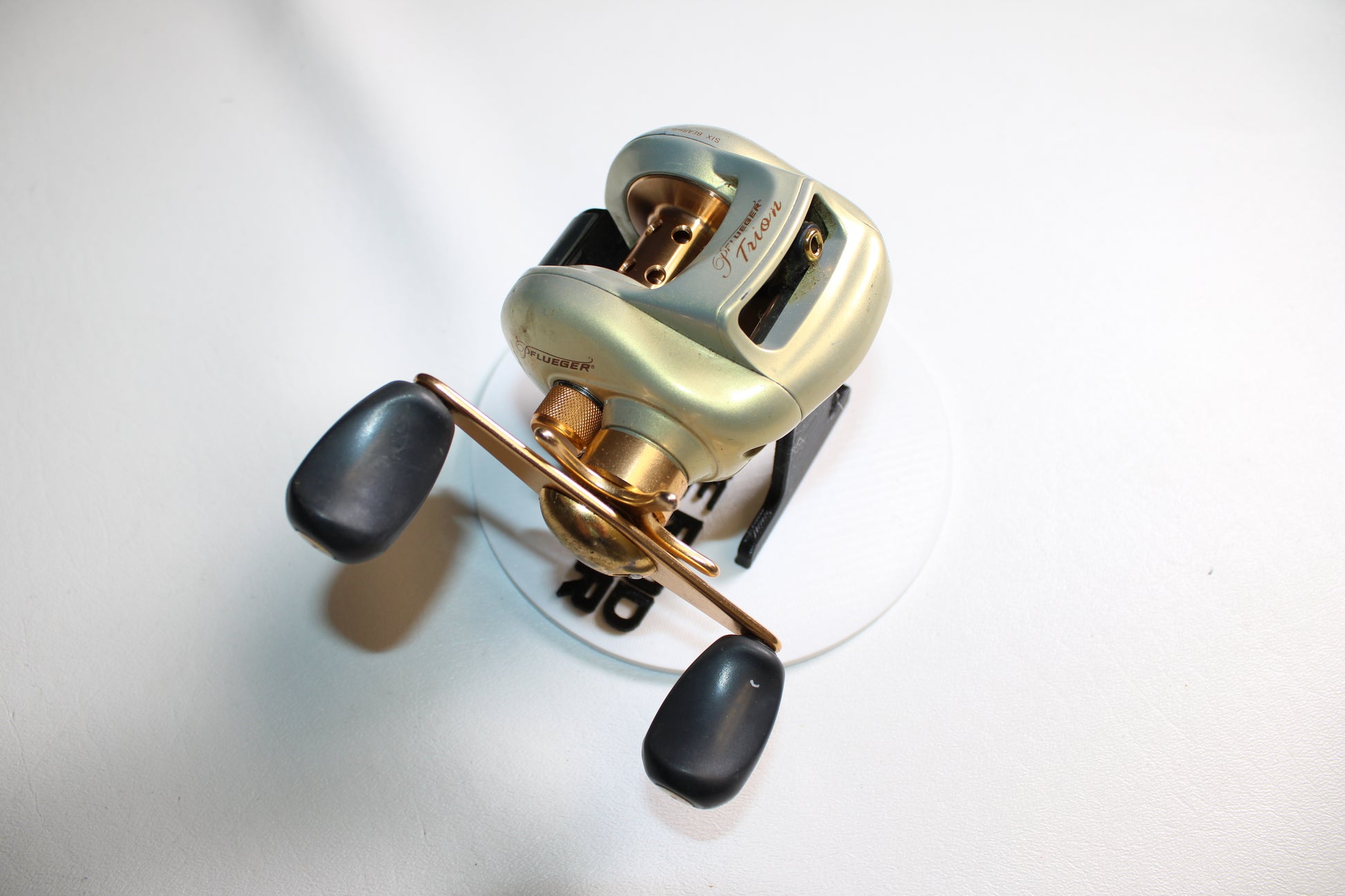A close-up of a Pflueger Trion LP 6.3:1 Gear Ratio Casting Reel in good used condition, featuring black handles and placed on a white surface.