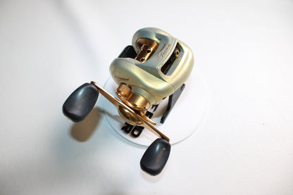 A close-up of a Pflueger Trion LP 6.3:1 Gear Ratio Casting Reel in good used condition, featuring black handles and placed on a white surface.