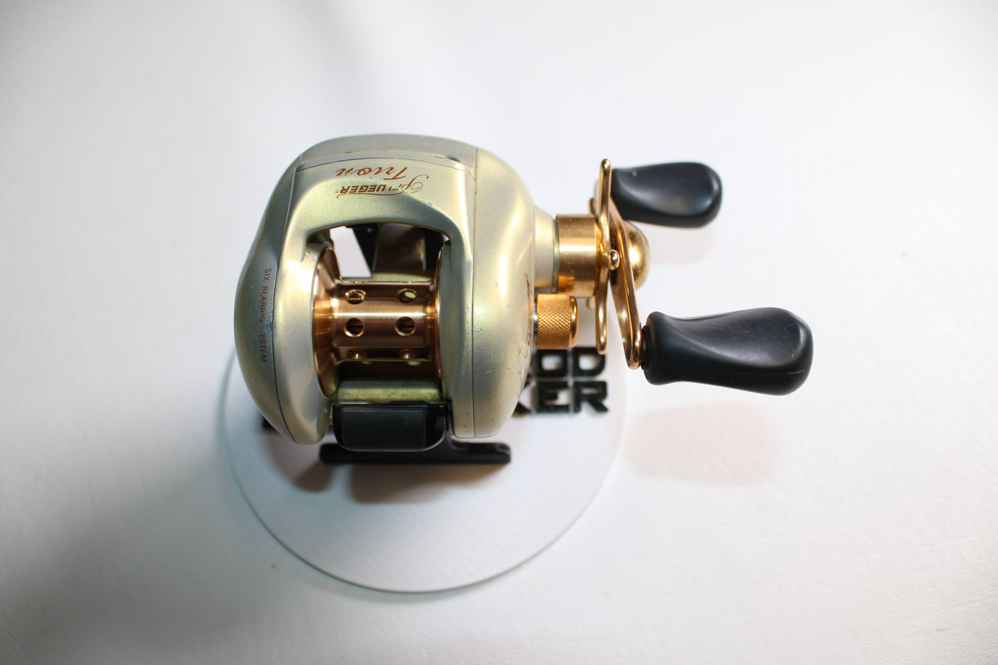 A Pflueger Trion LP 6.3:1 Gear Ratio casting reel in good used condition, featuring gold and black dual handles, is displayed on a white surface and photographed from above.