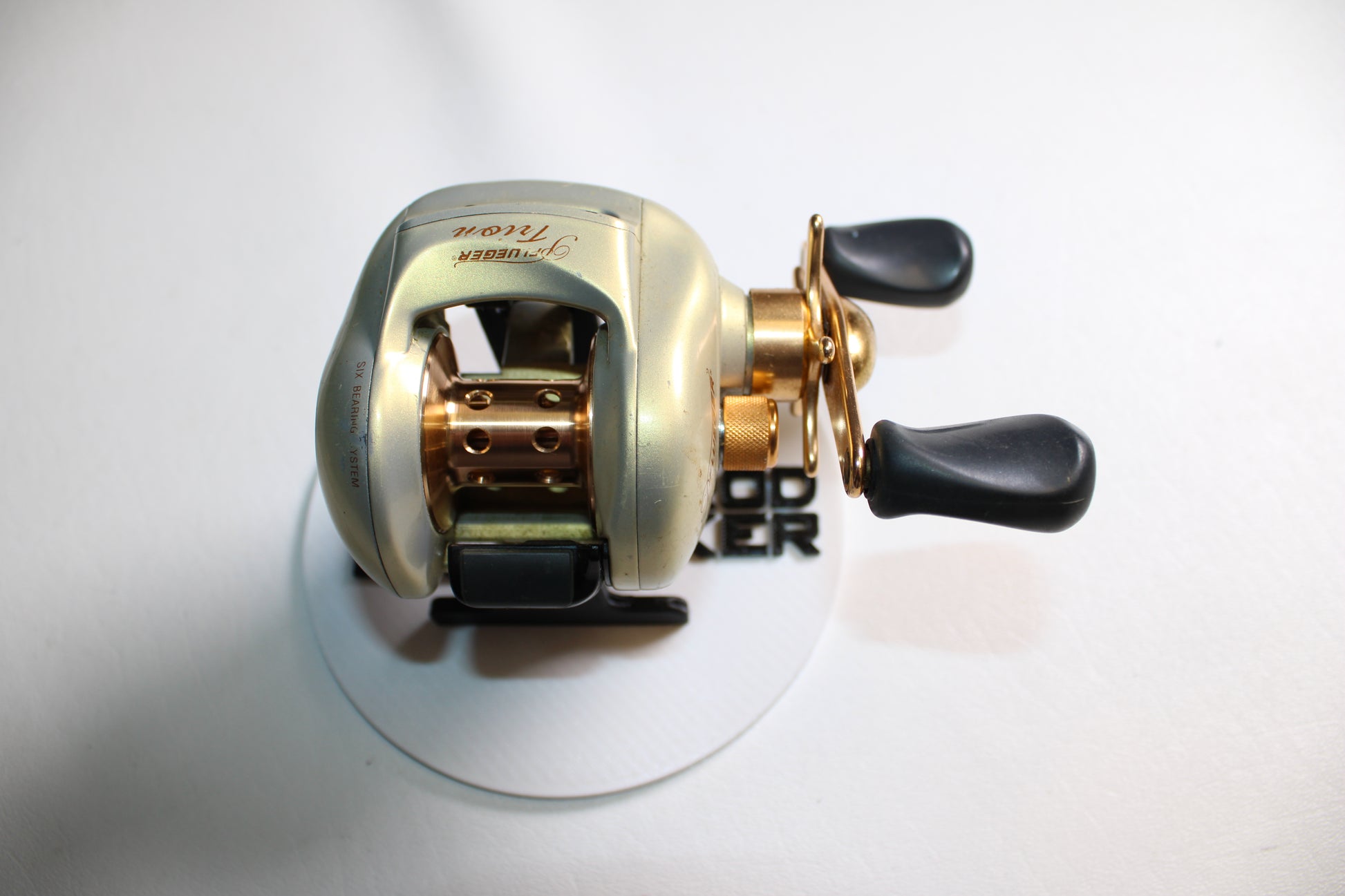 A Pflueger Trion LP 6.3:1 Gear Ratio casting reel in good used condition, featuring gold and black dual handles, is displayed on a white surface and photographed from above.