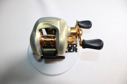 A Pflueger Trion LP 6.3:1 Gear Ratio casting reel in good used condition, featuring gold and black dual handles, is displayed on a white surface and photographed from above.