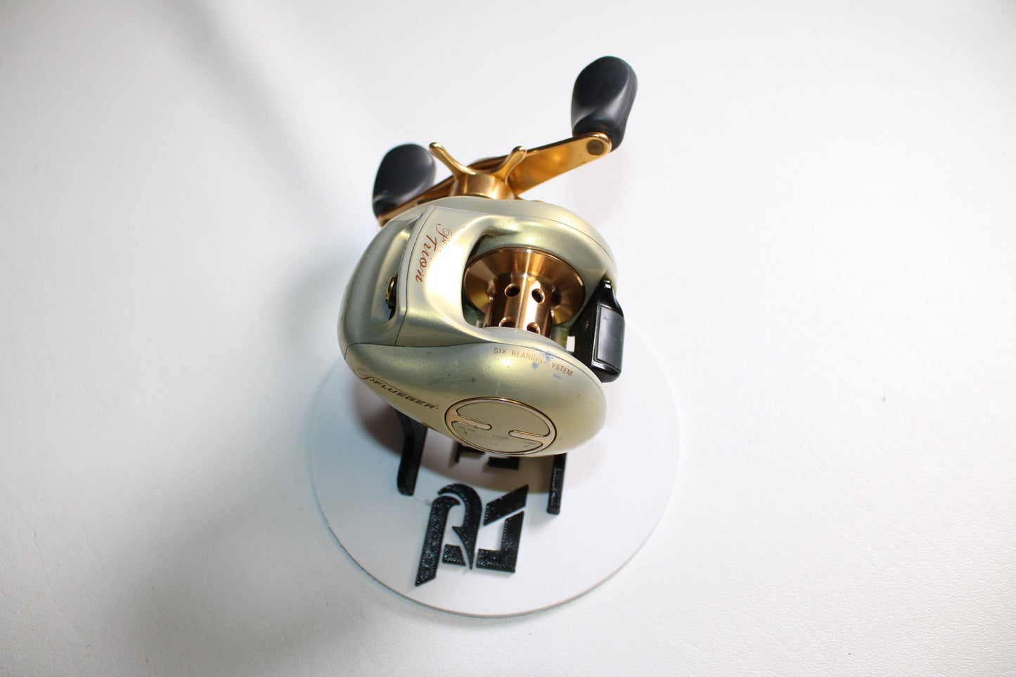 A Pflueger Trion LP 6.3:1 Gear Ratio Casting Reel in good used condition rests on a white circular surface with black letters, against a white background.