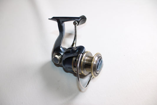 A close-up of the Pflueger President 6930 Used Spinning Reel by Shimano in very good condition, highlighting its metal spool, handle, and bail arm on a white surface.
