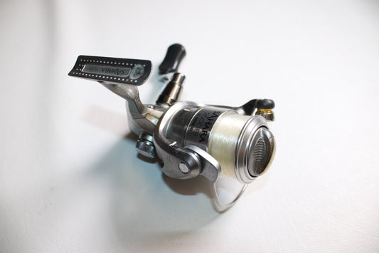 A close-up of a Shimano Sahara 2500FB used spinning reel in fair condition, featuring white fishing line on the spool and placed on a plain white surface.