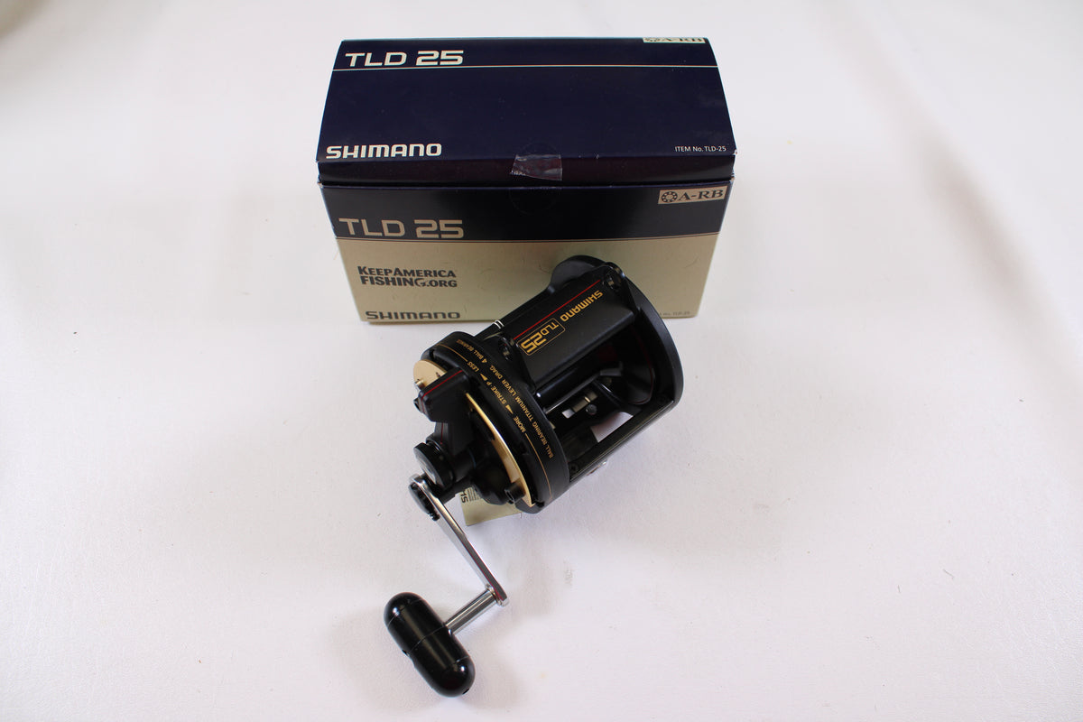 A Shimano TLD TLD25 Used Conventional Reel in mint condition is shown in front of its original blue and beige branded box on a white surface. The black reel features gold accents and a handle.