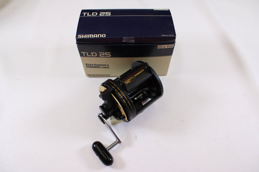 A Shimano TLD TLD25 Used Conventional Reel in mint condition is shown in front of its original blue and beige branded box on a white surface. The black reel features gold accents and a handle.