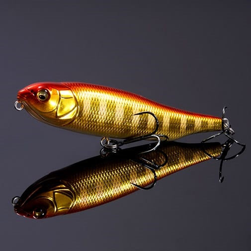A Megabass Giant Dog-X (SP-C) M Akakin pencil bait in shiny gold and red, featuring two treble hooks, is displayed on a reflective black surface.