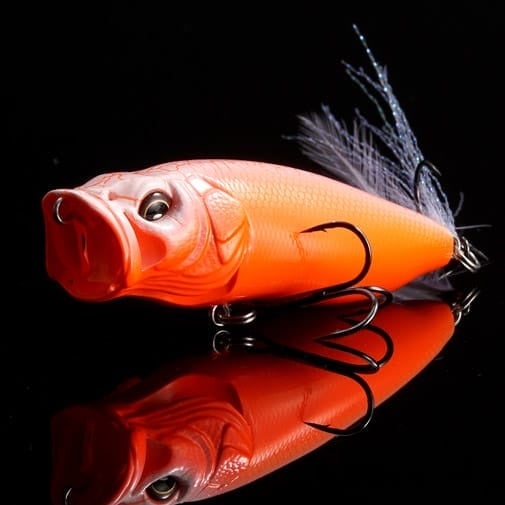 A Megabass PopMax (SP-C) Secret V-Ore fishing lure by Megabass, with vivid scales, black eyes, and treble hooks, rests on a glossy black surface, its feathered tail slightly raised.