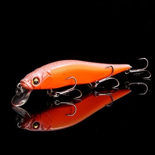 The Megabass Ito Vision 110 Jr. Jerkbait (SP-C) Secret V-Ore, a fish-shaped orange lure with three treble hooks and a clear plastic lip, is displayed on a reflective black surface.