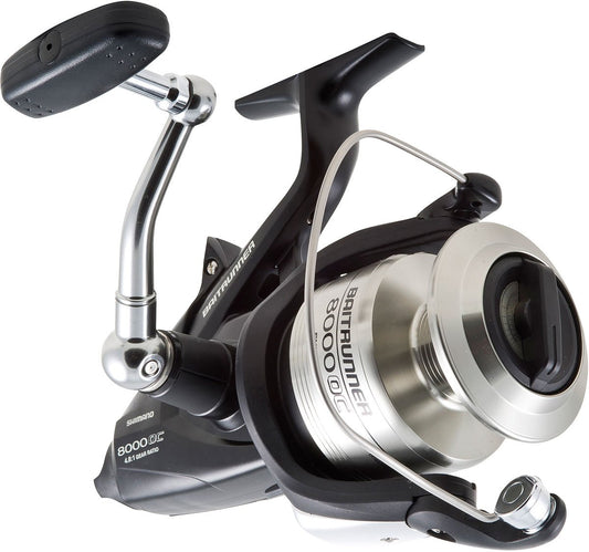 A close-up of the Shimano SHIMANO Baitrunner OC BTR8000OC spinning reel highlights its handle, spool, and metal bail arm, featuring a sleek black and silver finish.
