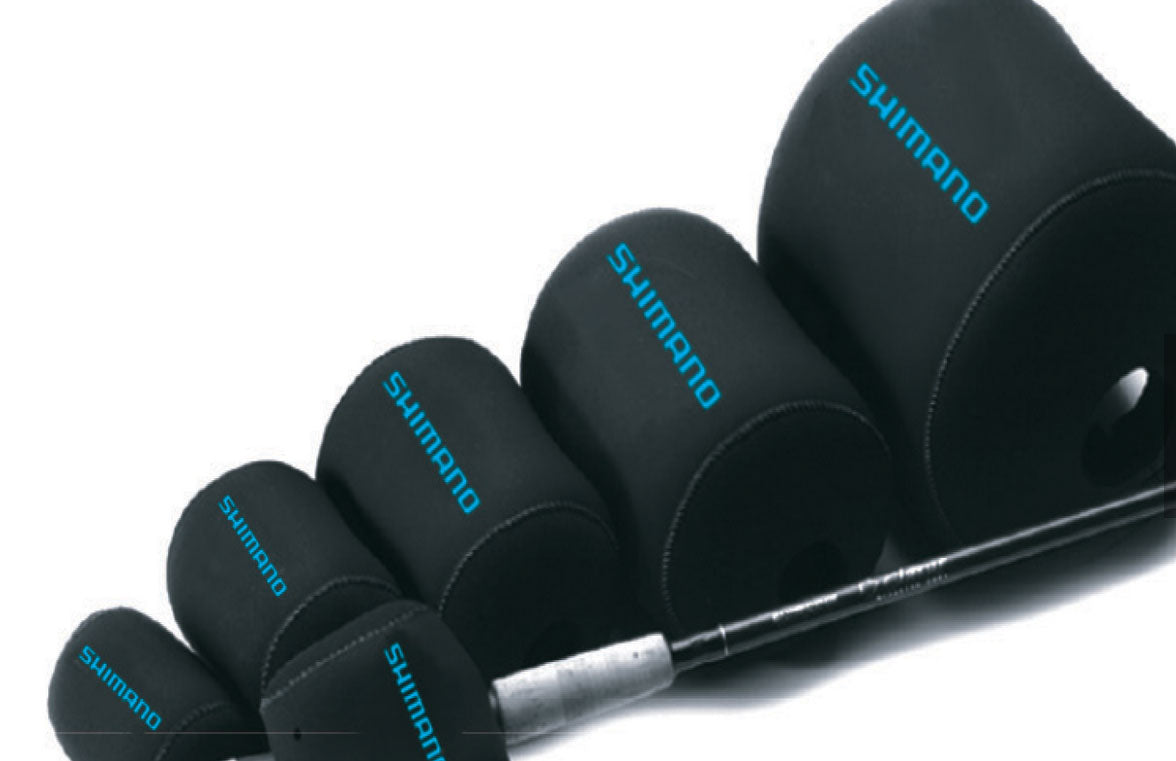 Five Shimano Neoprene Casting Reel Covers in black, each labeled "SHIMANO" in blue, are arranged by size, with a fishing rod and baitcasting reel partially visible through the smallest cover.