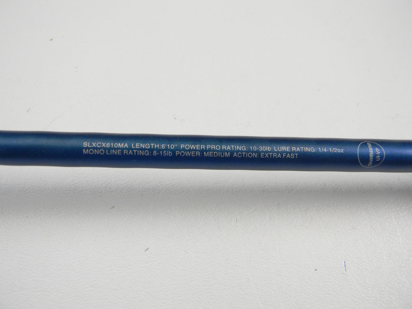 A close-up of a blue Shimano SLX SLXCX610MA 6'10" Medium used casting rod in excellent condition, showing white specs text. Ideal for those seeking used Shimano gear or a rod and reel trade-in. The rod rests on a white surface.