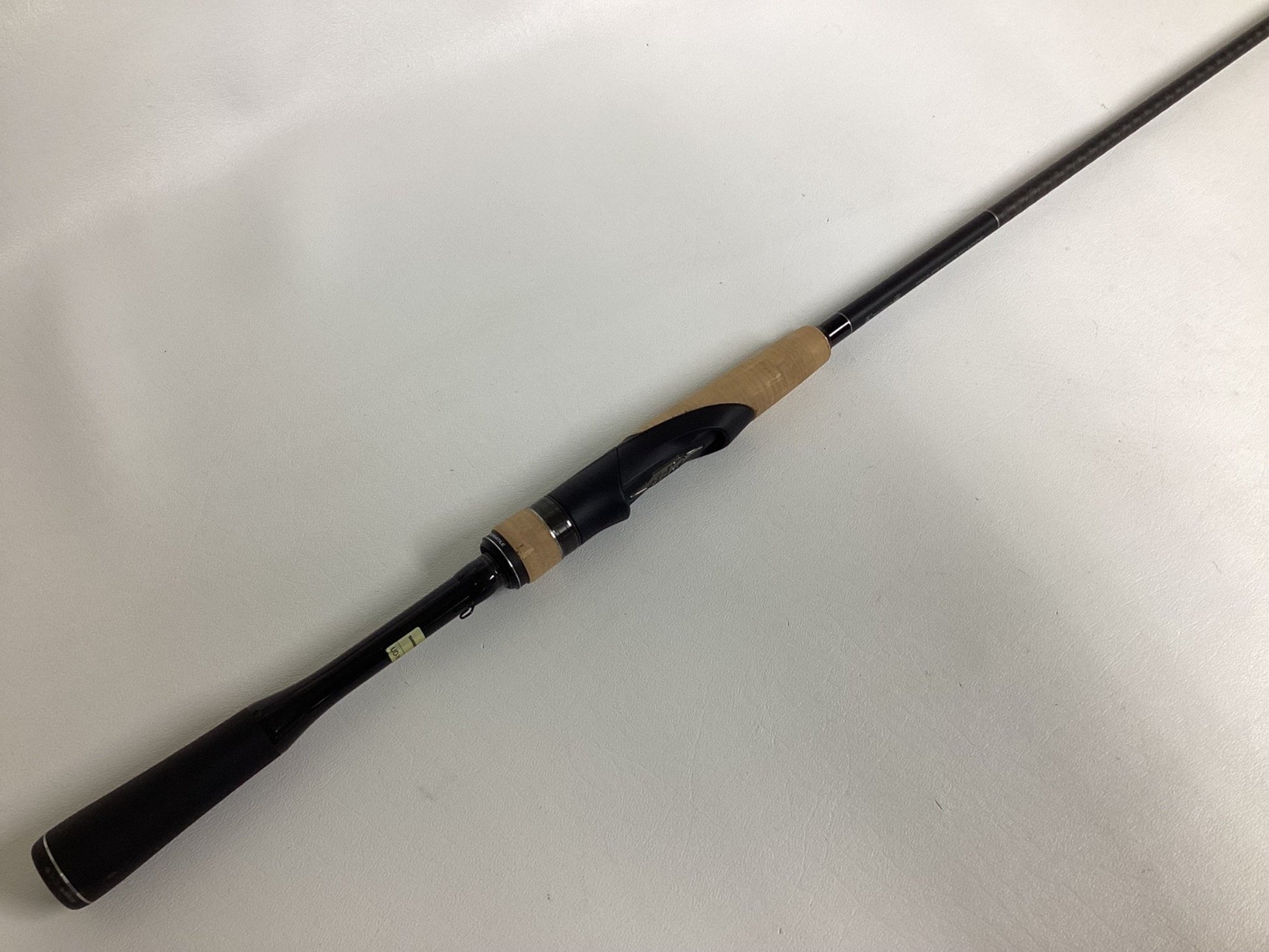 The Shimano Expride B EXS70LB, a used spinning rod in excellent condition, features a black handle with a light brown cork grip. Placed diagonally on a white surface, the black reel seat is visible as the rod extends out of frame.