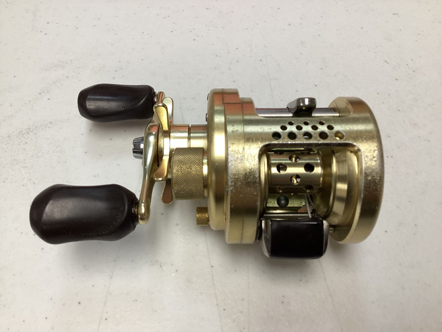 A Shimano Calcutta Conquest 101A 5.6:1 LH used baitcasting reel in fair condition, gold-colored with two black handles and perforated spool housing, on a light gray surface; eligible for our Rod and Reel Trade-In Program for store credit.