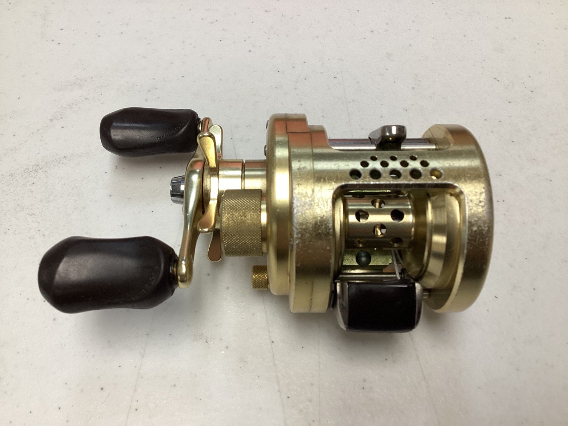 A Shimano Calcutta Conquest 101A 5.6:1 LH used baitcasting reel in fair condition, gold-colored with two black handles and perforated spool housing, on a light gray surface; eligible for our Rod and Reel Trade-In Program for store credit.