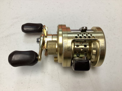 A Shimano Calcutta Conquest 101A 5.6:1 LH used baitcasting reel in fair condition, gold-colored with two black handles and perforated spool housing, on a light gray surface; eligible for our Rod and Reel Trade-In Program for store credit.