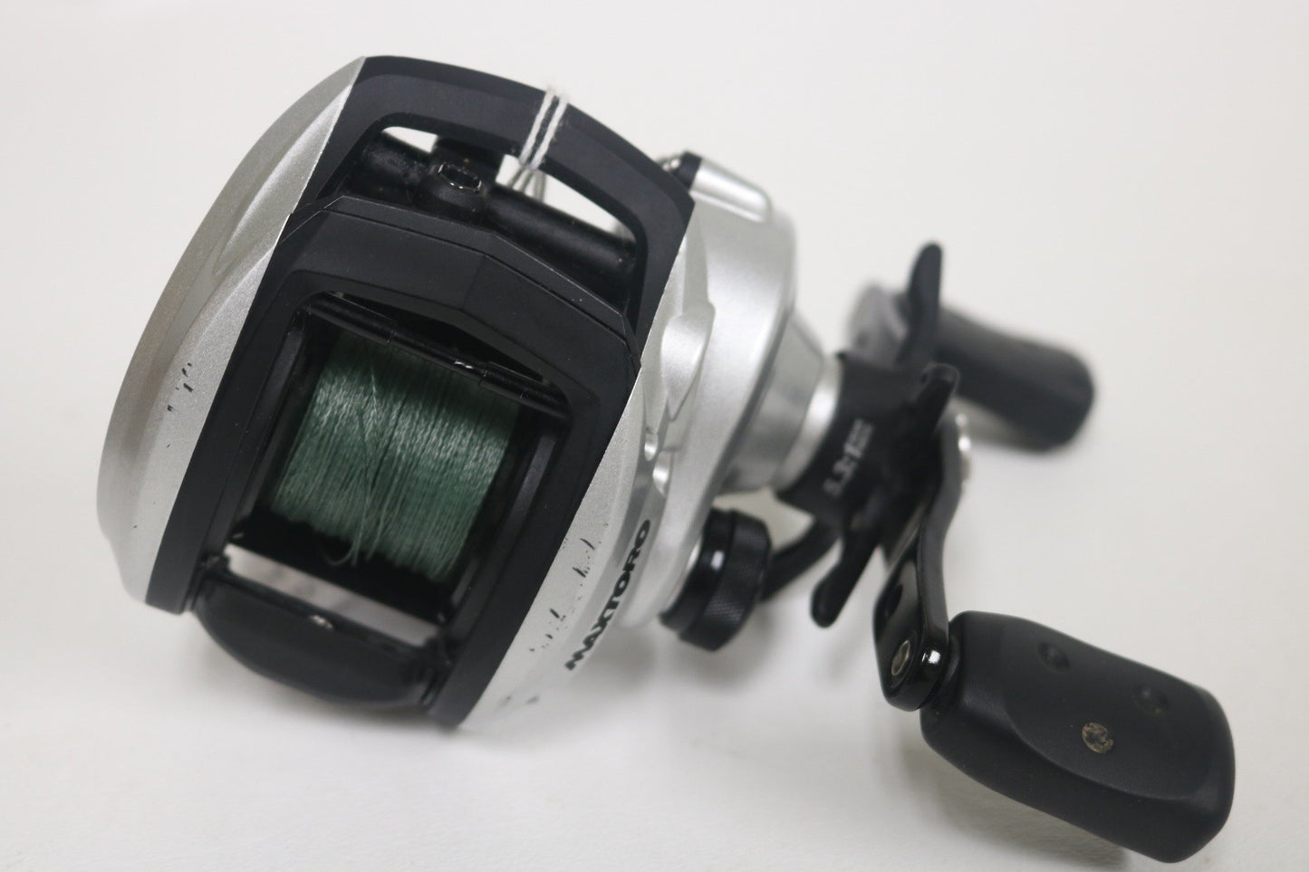 A close-up of an Abu Garcia MAXTORO 5.3:1 Casting Reel (Used - Very Good Condition) with green fishing line, silver and black body, and dual-handle grip, shown against a plain white background.