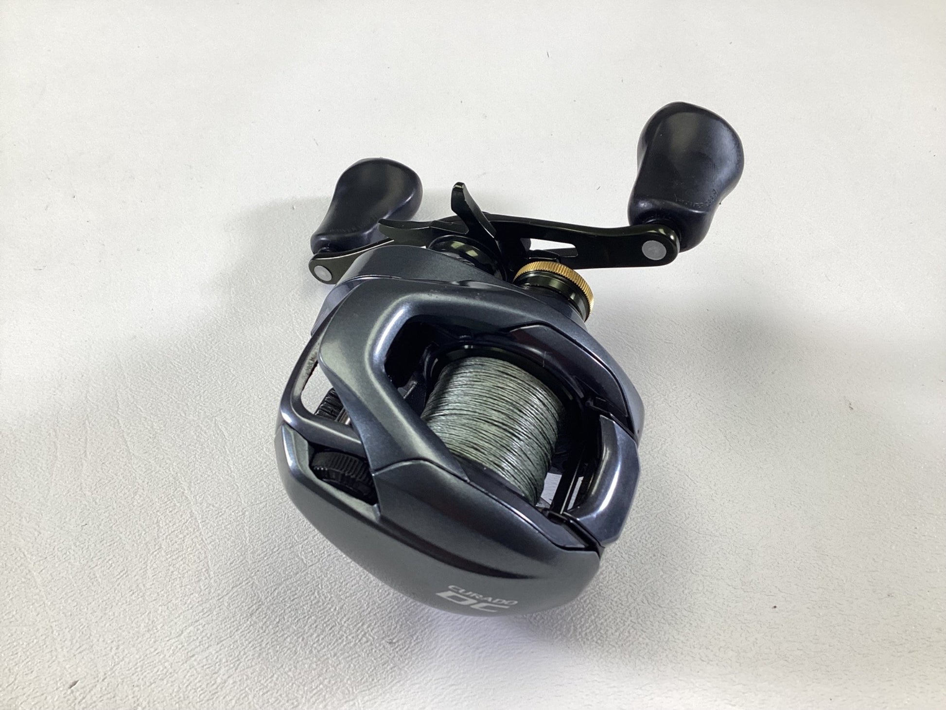 A Shimano Curado CUDC150XG used casting reel, featuring a sleek metallic finish with dual handles and dark-colored accents, is displayed on a light gray surface. This compact design is available through our Rod and Reel Trade-In program, providing anglers the opportunity to upgrade their gear while receiving store credit.